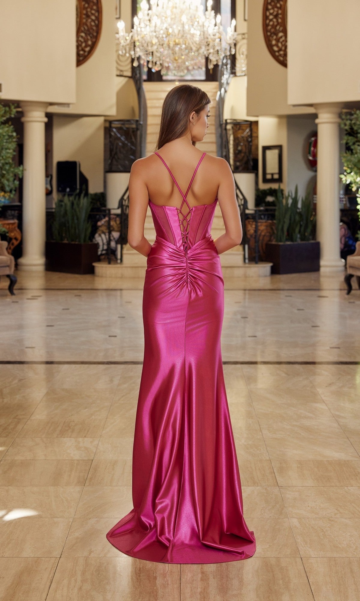 A woman wears the Nox Anabel R1813 long prom dress—a shiny pink gown with crisscross straps and a thigh-high slit—standing in an elegant lobby with chandeliers and columns.
